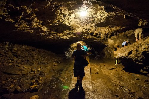 A person walks through a dimly lit cave, with rocky walls and a wet floor, while others explore in the background.