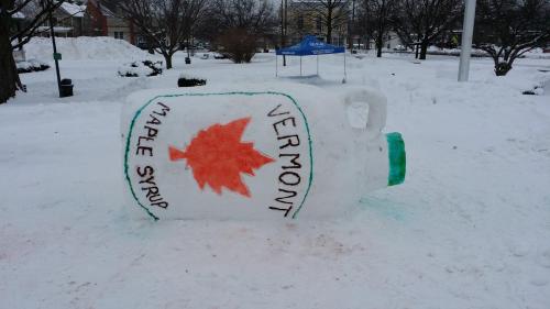 A large snow sculpture of a maple syrup bottle, labeled "Vermont," in a snowy park setting.