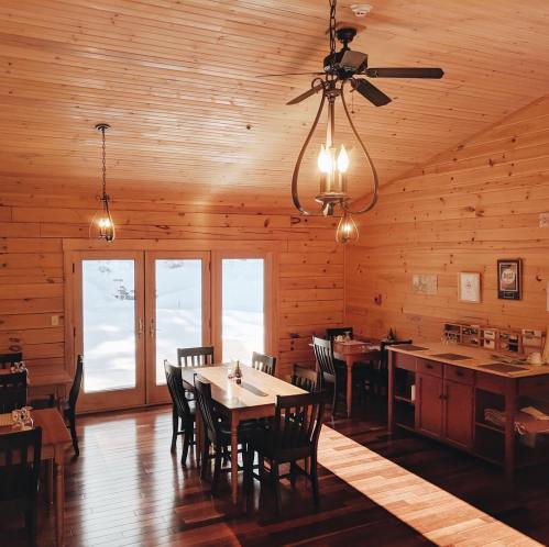 Cozy wooden dining area with tables, chairs, and large windows overlooking a snowy landscape. Warm lighting enhances the ambiance.