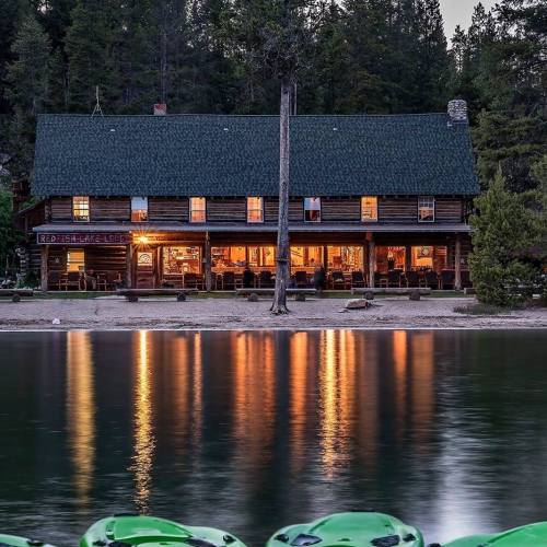 A rustic lodge by a lake, illuminated at dusk, with reflections on the water and surrounded by trees.