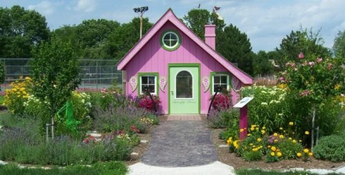 A charming pink cottage surrounded by colorful flowers and greenery, with a stone pathway leading to the entrance.