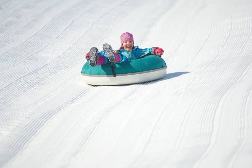 A child in a colorful winter outfit joyfully sleds down a snowy slope on a green inner tube.