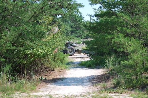 A sandy path through greenery leads to a military vehicle partially obscured by trees.