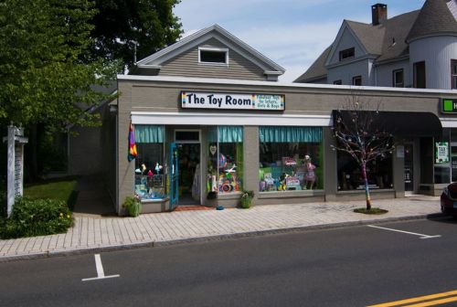 A colorful toy store called "The Toy Room" with large windows displaying various toys, set on a sunny street.