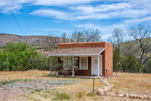 A small adobe house with a porch, surrounded by dry grass and trees, set against a backdrop of rolling hills and blue sky.