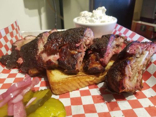 A plate of smoked ribs on toast, garnished with pickles, onions, and a bowl of creamy coleslaw.