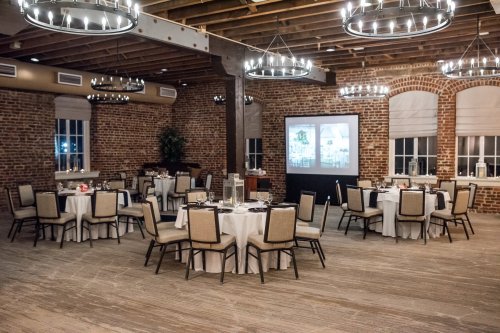 Elegant dining room with round tables, white tablecloths, and exposed brick walls, featuring chandeliers and a projector screen.