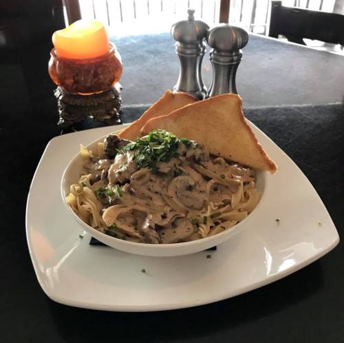 A bowl of creamy mushroom pasta topped with herbs, accompanied by two slices of toasted bread and a candle in the background.