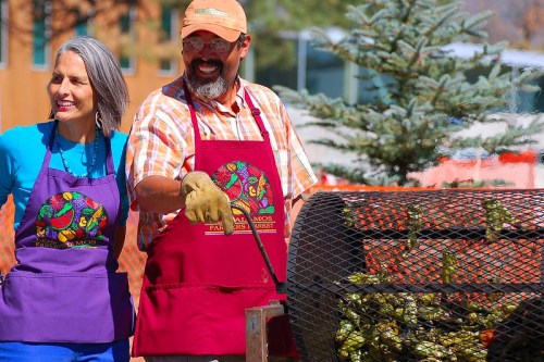 A man and woman in aprons smile while grilling vegetables at a farmers market. Trees and a building are in the background.
