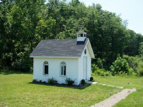 A small white house with a bell tower, surrounded by greenery and a gravel path leading to it.