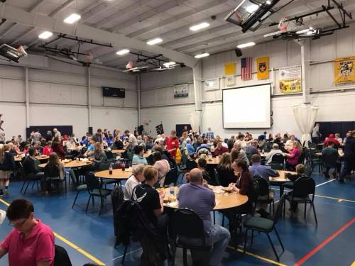 A large group of people dining in a gymnasium, with tables set up and a projector screen in the background.