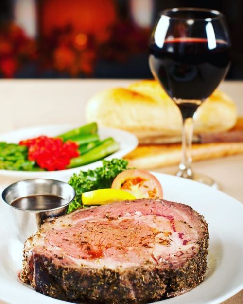 A plate of prime rib with herbs, served with asparagus, a slice of tomato, and a glass of red wine in the background.