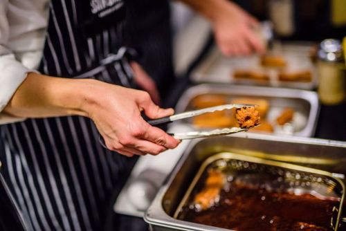 A chef uses tongs to lift a piece of fried food from a tray, with another person preparing food in the background.