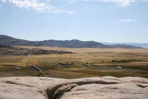 A vast, open landscape with rolling hills, a river, and a distant mountain range under a clear blue sky.
