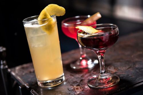 Three cocktails on a bar: a tall drink with a lemon twist, and two shorter drinks with garnishes in coupe glasses.