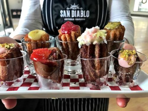 A platter of mini churros topped with various colorful sauces, served in small cups on a checkered tray.