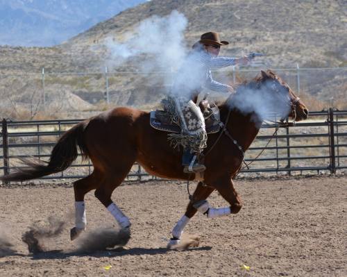A cowboy on a brown horse fires a gun while riding in a dusty arena, with smoke trailing from the weapon.