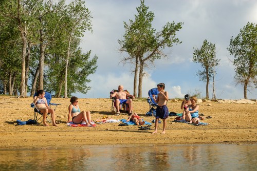 A group of people relaxes on a sandy beach by the water, enjoying a sunny day with trees in the background.