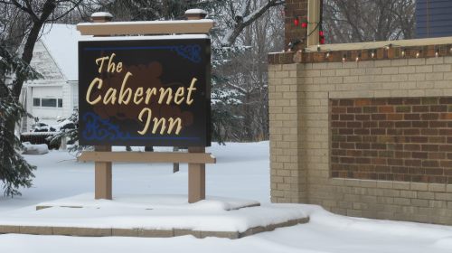 Sign for "The Cabernet Inn" in a snowy landscape, with decorative elements and a brick building in the background.