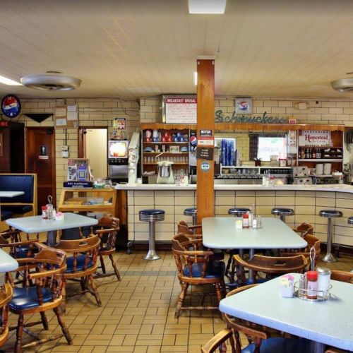 A cozy diner interior with wooden tables, chairs, a counter, and a vintage decor featuring a soda fountain and menu board.