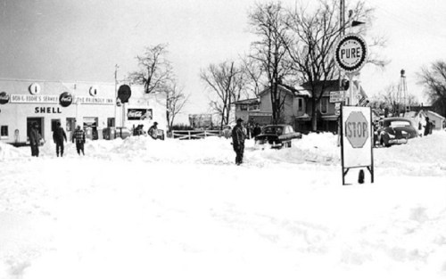 Historic snowy scene featuring a gas station, vintage cars, and people navigating deep snow.