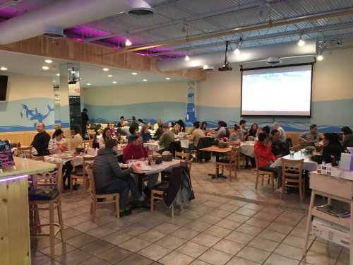 A busy restaurant interior with diners at tables, a large screen displaying a game, and a coastal-themed decor.