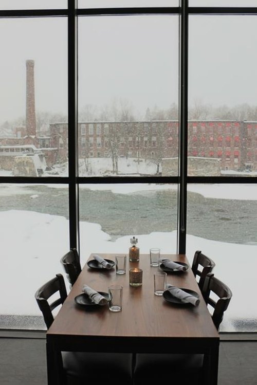 A cozy dining table set for four, with a snowy river view and a brick building in the background.