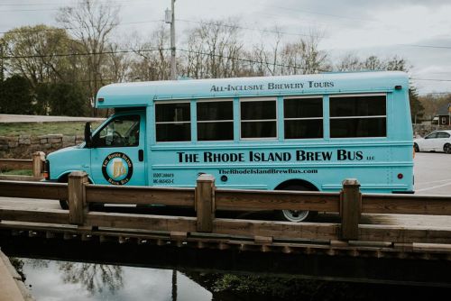 A turquoise bus labeled "The Rhode Island Brew Bus" parked near a wooden bridge, promoting brewery tours.
