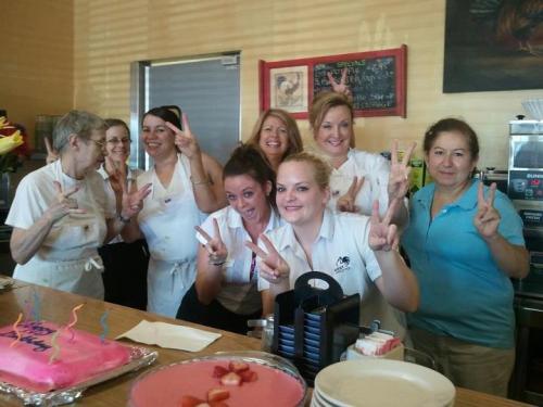 A group of smiling women in a café, posing with peace signs, surrounded by cakes and a cheerful atmosphere.