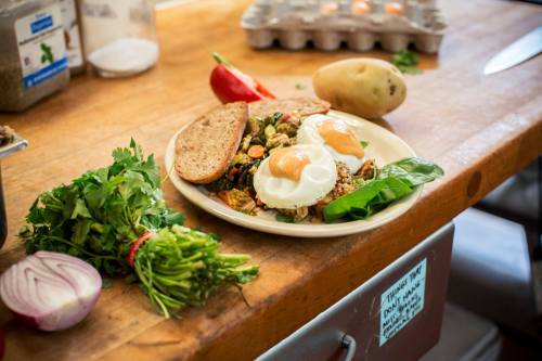 A plate with two poached eggs on a vegetable mix, served with whole grain bread, herbs, and a potato on a wooden table.