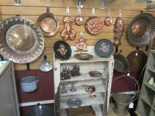 A display of vintage copper cookware and decorative items on a wall, with a rustic shelf holding various antiques.