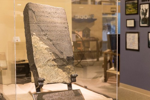 A large stone tablet with ancient inscriptions displayed in a glass case in a museum setting.