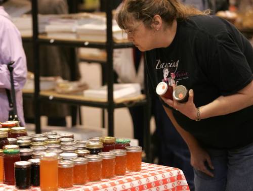 A woman examines jars of colorful jams and jellies displayed on a table at a market.
