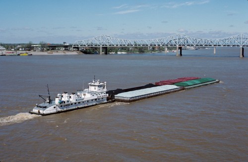 A barge navigates a river, transporting cargo beneath a bridge on a clear day.