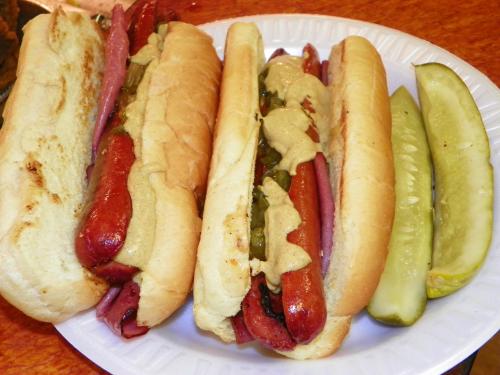 Three hot dogs in buns topped with mustard and pickles on the side, served on a white plate.