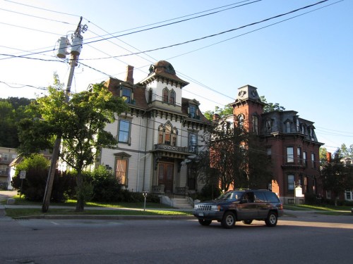 A vintage-style house with ornate architecture next to a red brick building, with a car passing by on the street.