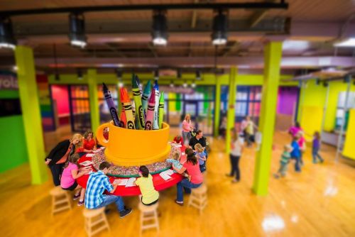 A colorful art room with a large crayon cup centerpiece and children engaged in creative activities around a circular table.