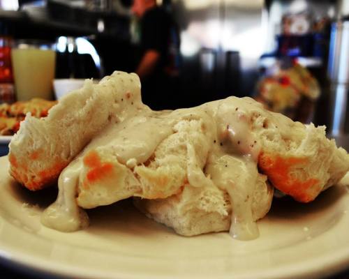 A plate of fluffy biscuits smothered in creamy white gravy, with a blurred kitchen background.