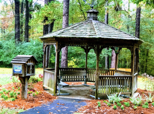 A wooden gazebo surrounded by trees, with a small mailbox nearby and flower beds in the foreground.