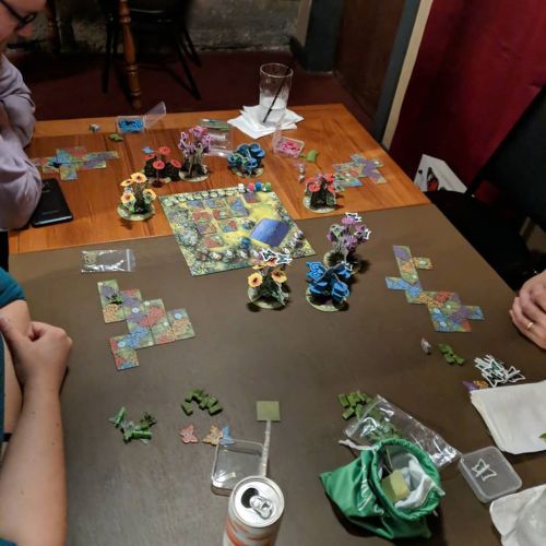 A tabletop game in progress with colorful pieces, flowers, and a game board on a wooden table surrounded by players.