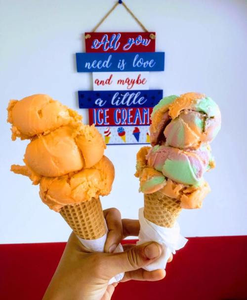 A hand holds two ice cream cones: one with orange scoops and the other with colorful mint and orange scoops, against a wall sign.