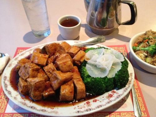 A plate of glazed tofu cubes served with broccoli and a radish garnish, alongside tea and a bowl of fried rice.