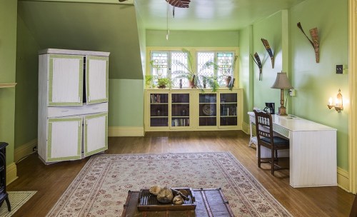 A bright green room with a white cabinet, a desk, bookshelves, and plants by the window, featuring wooden flooring and a rug.
