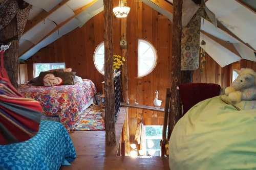Cozy wooden loft with colorful bedding, a round window, and a whimsical decor featuring a stuffed animal and flowers.