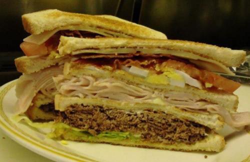A towering sandwich with layers of turkey, ham, bacon, lettuce, tomato, and mayonnaise between toasted bread.