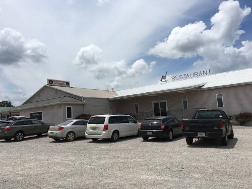 A restaurant building with a gravel parking lot, featuring several parked cars and a cloudy sky above.