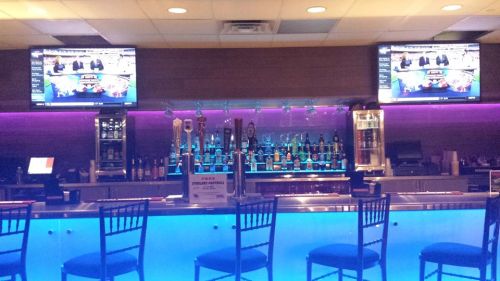 A modern bar with illuminated blue lighting, featuring taps and screens displaying news or sports.