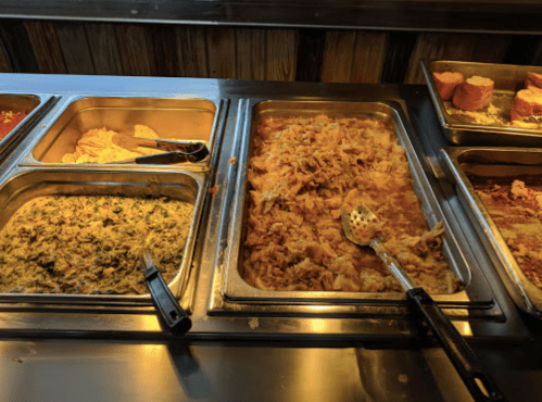 A buffet display with various dishes, including green vegetables, rice, and fried snacks in metal trays.