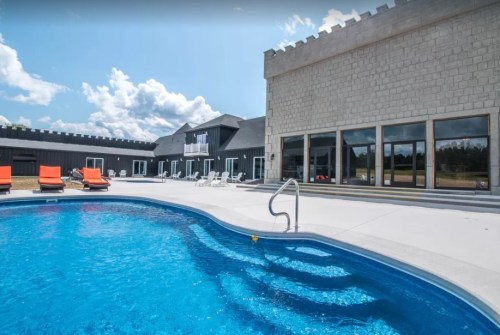 A sunny pool area with orange lounge chairs, surrounded by a modern building and blue sky with clouds.