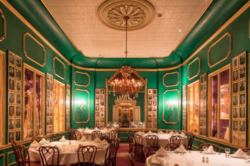 Elegant restaurant interior with green walls, framed photos, and a chandelier, featuring round tables set for dining.